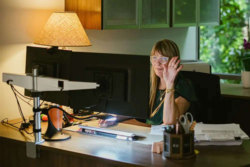 A woman sitting at her computer in a dark lit office waving and smiling