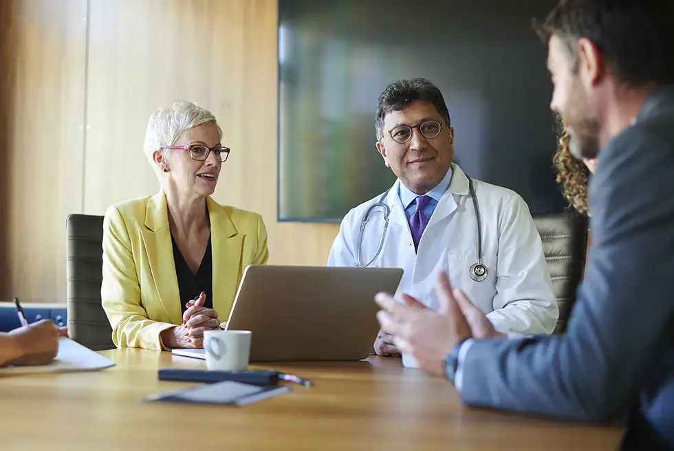 Businesswoman in yellow suit with a laptop in front of her sitting next to a doctor talking to other people at a table in an office