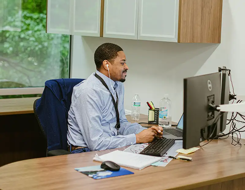 A man with earbuds in sitting at his work desk typing on his computer 