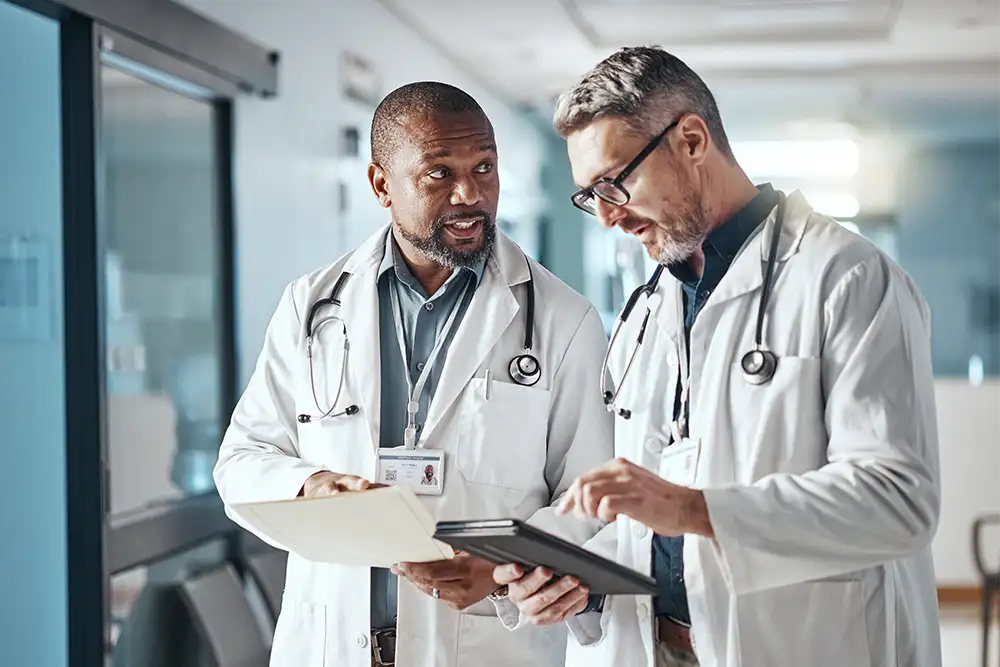Two male physicians standing in a hallway talking and looking at paperwork