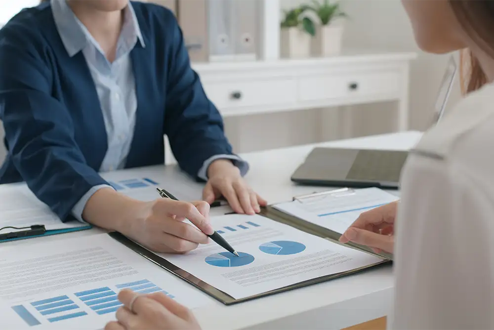 Close up of a business person holding a pen over charts and graphs on a desk while another person sits on the opposite side