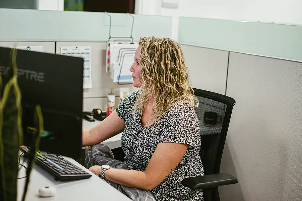 A blonde woman sitting at her desk typing on her computer in an office cubicle