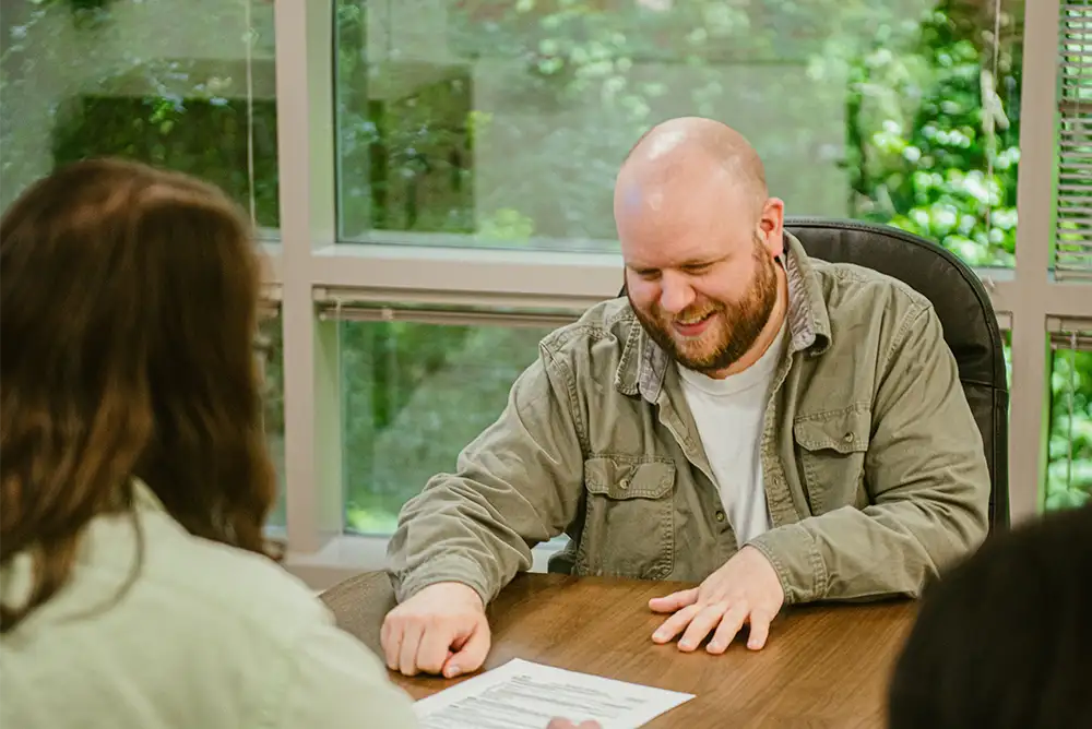 A man in a green shirt smiling and looking at a sheet of paper while he sits at an office table