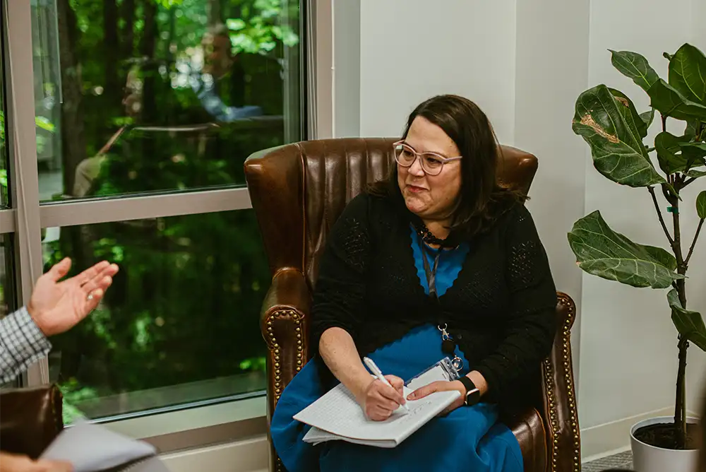 Woman with dark hair and glasses sitting in a brown leather armchair with a notebook on her lap