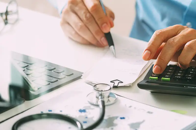 A close up of a physicians hand tapping on a calculator and writing on paper with a stethoscope on the table 