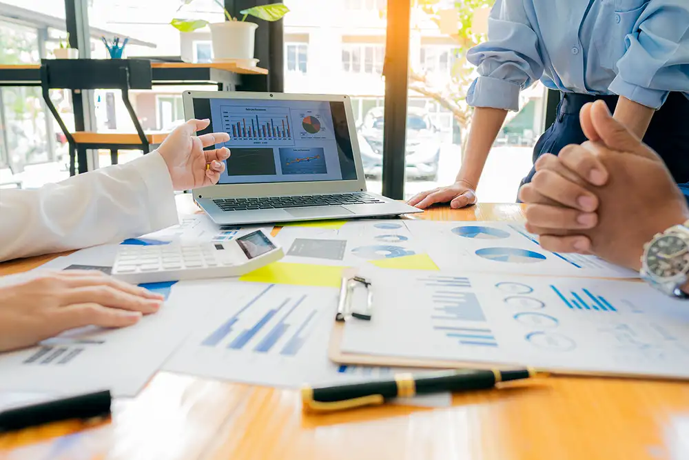 Close up of a table filled with graphs, clipboards, pens, a calculator and a laptop