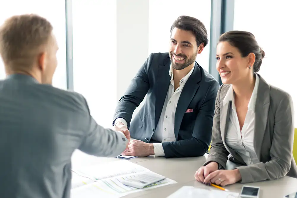 A businessman smiling and shaking another mans hand while a businesswoman smiles next to him