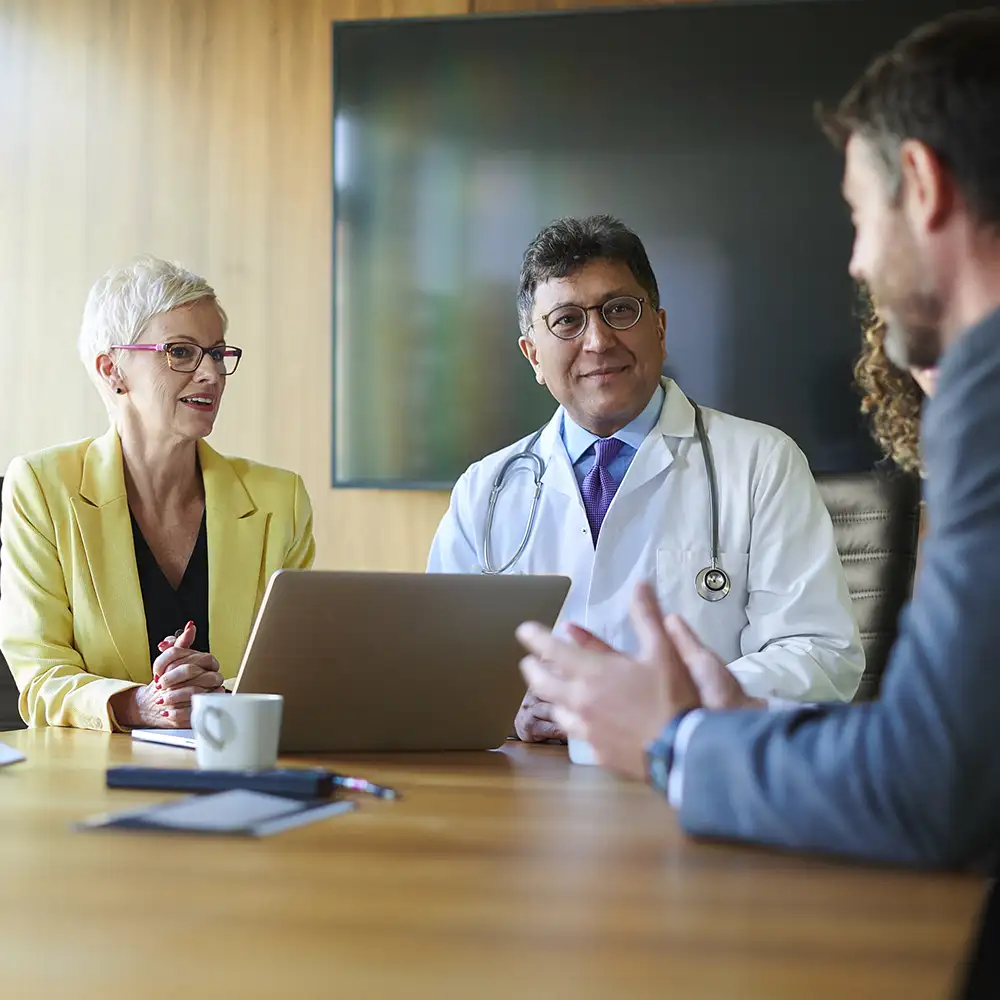 Businesswoman in yellow suit with a laptop in front of her sitting next to a doctor talking to other people at a table in an office