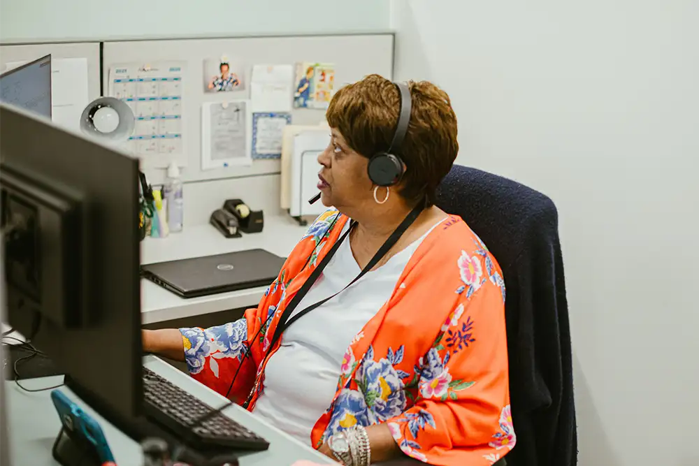 A woman in an orange sweater wearing a headset and looking at her computer screen while sitting in her work cubicle