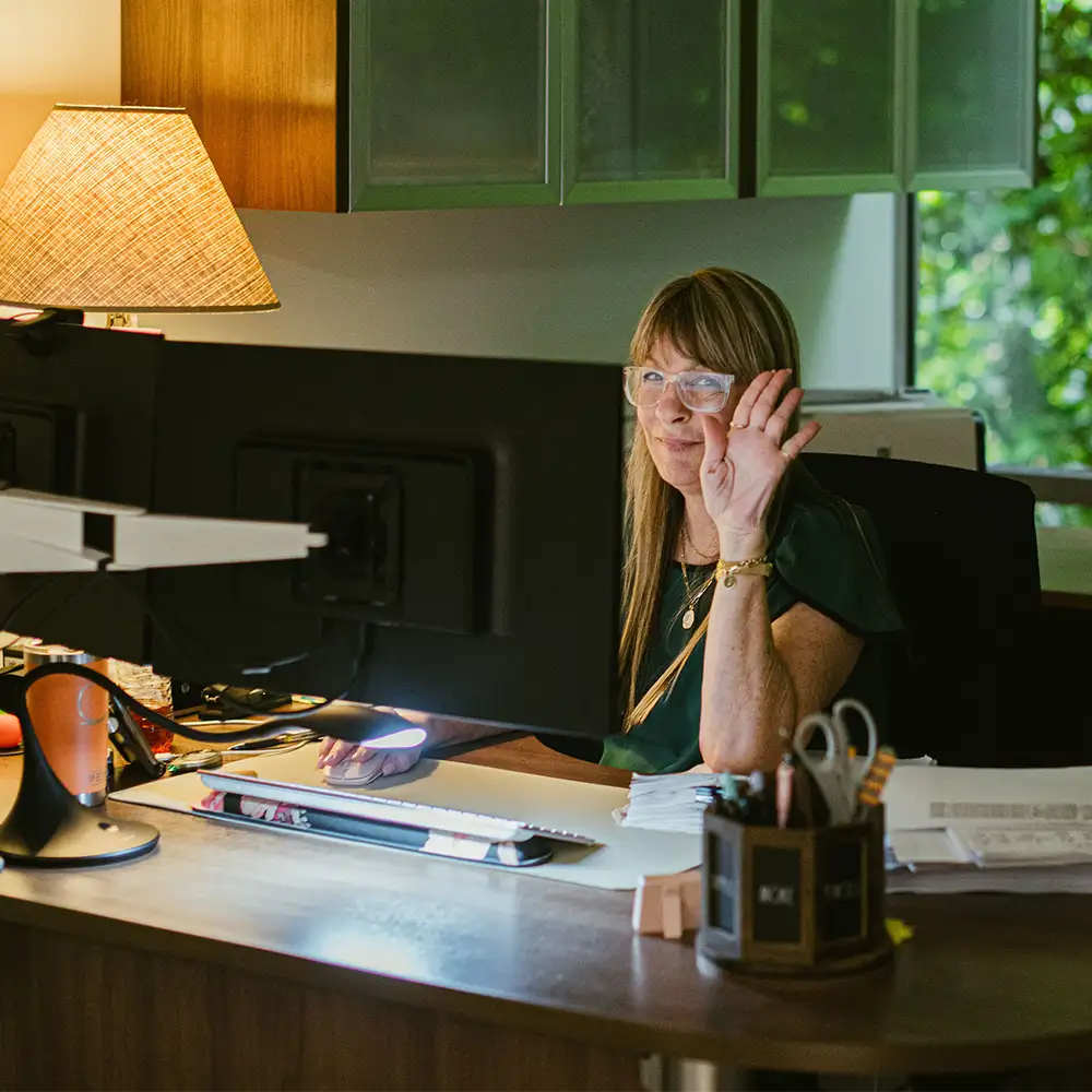 A woman sitting at her computer in a dark lit office waving and smiling