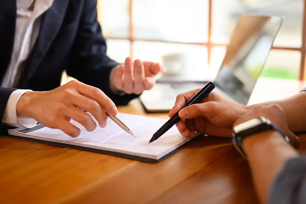 A close up of two peoples hands holding pens hovering over a document on a desk