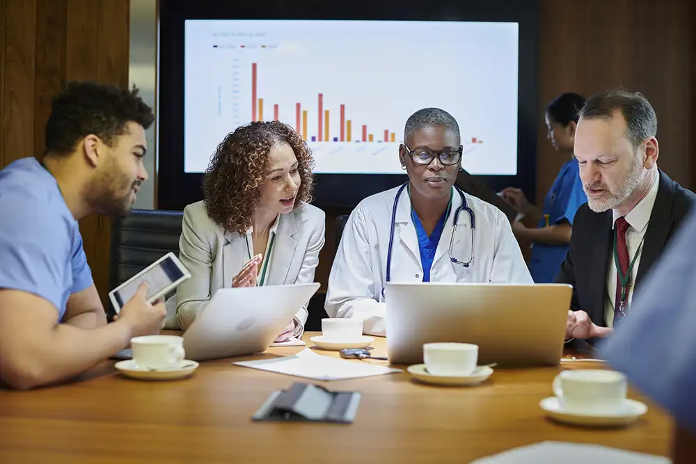 Physicians and business people sitting at a table with a screen displaying bar graphs behind them