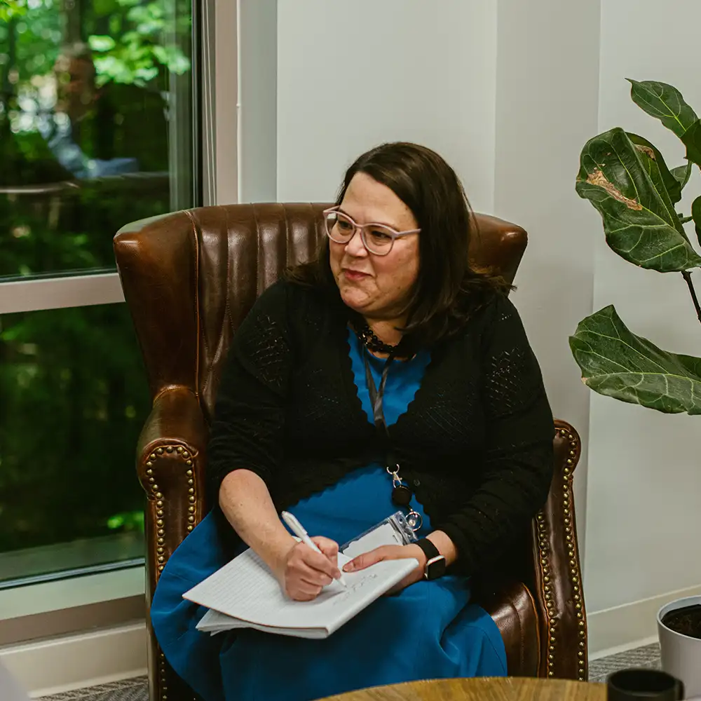 Woman with dark hair and glasses sitting in a brown leather armchair with a notebook on her lap