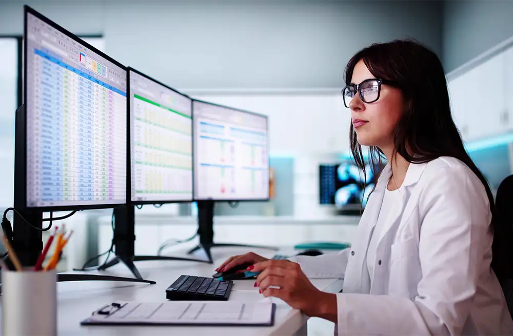 A woman with glasses sitting at her desk looking at 3 computer screens