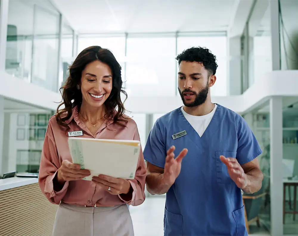 A young male physician talking and walking next to a young businesswoman in a bright hallway