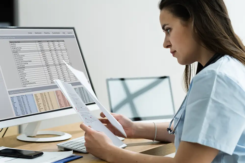 A woman sitting at her desk looking at papers monitors with spreadsheets on them