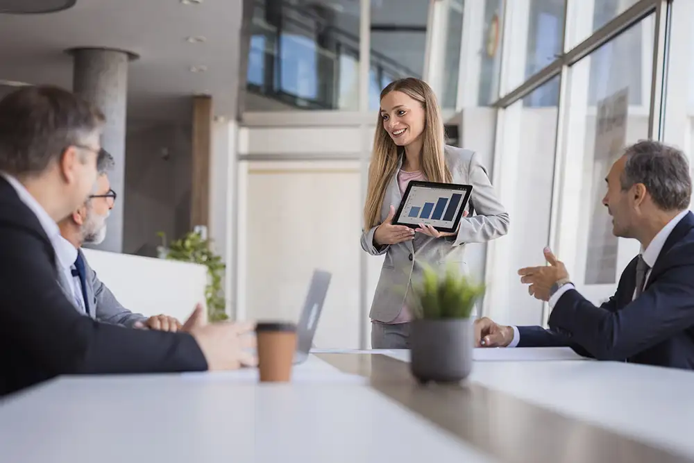 A businesswoman standing infront of a few businessmen sitting at a table showing them charts on a tablet