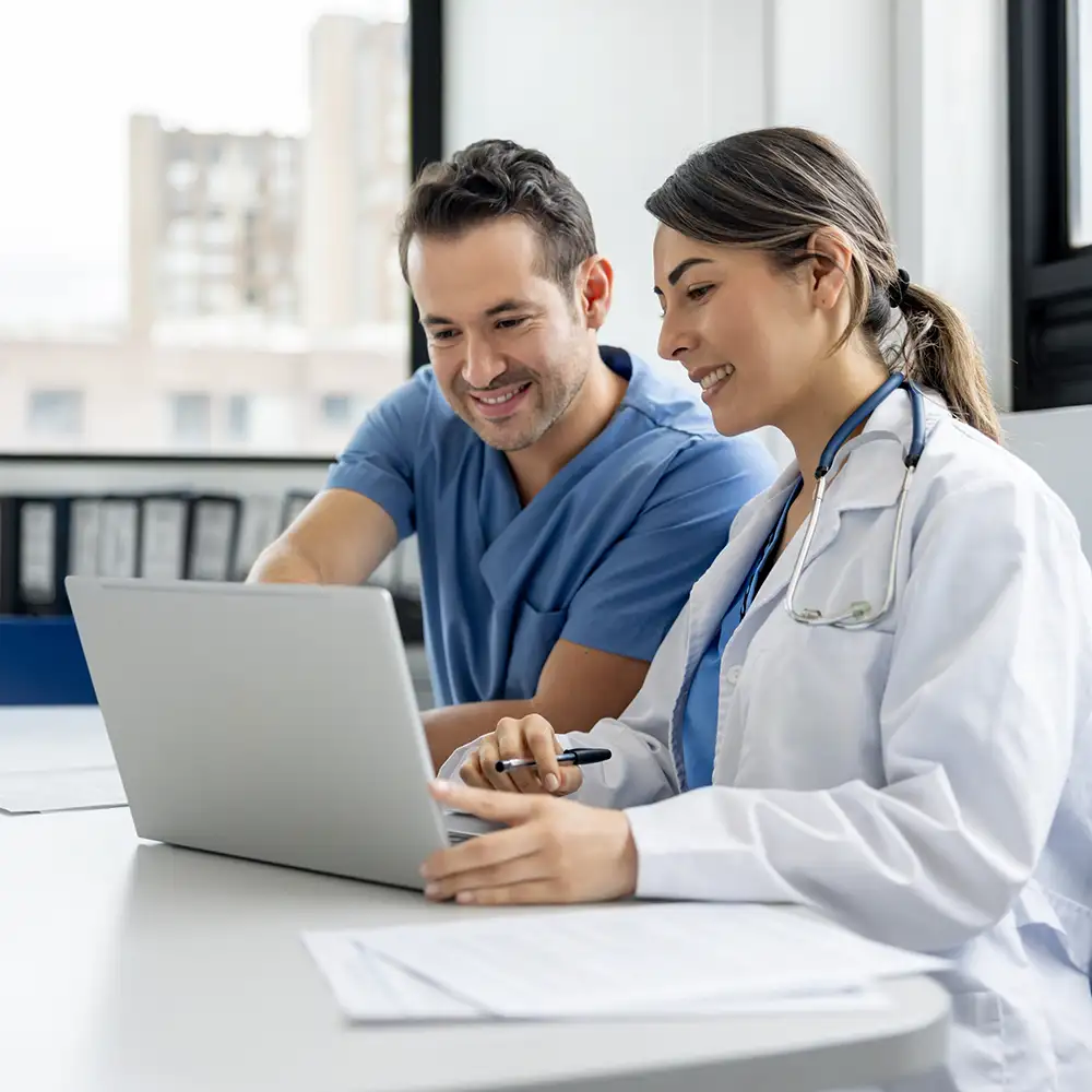 Two physicians sitting at a table smiling and looking at a laptop with papers in front of them