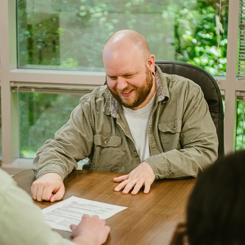 A man in a green shirt smiling and looking at a sheet of paper while he sits at an office table
