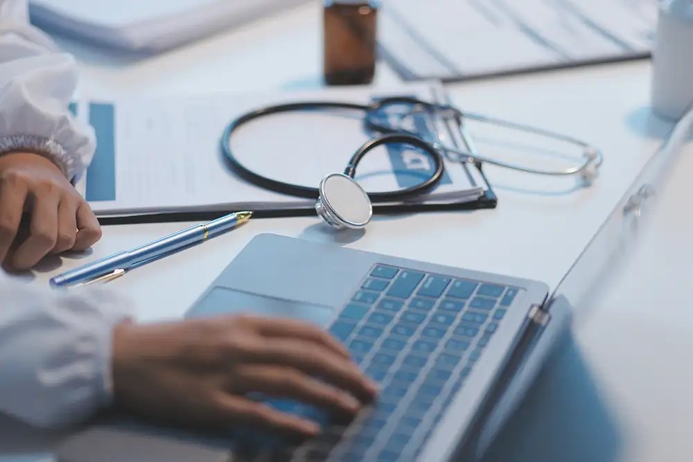 Close up view of a hand typing on a laptop with a stethoscope sitting off to the side