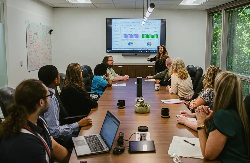 A team of business people sitting around a conference room table while a woman stands at the front presenting graphs and charts on a screen