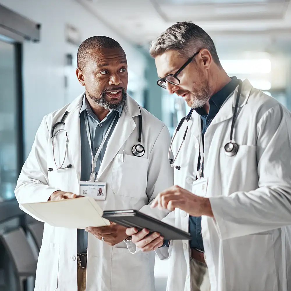 Two male physicians standing in a hallway talking and looking at paperwork