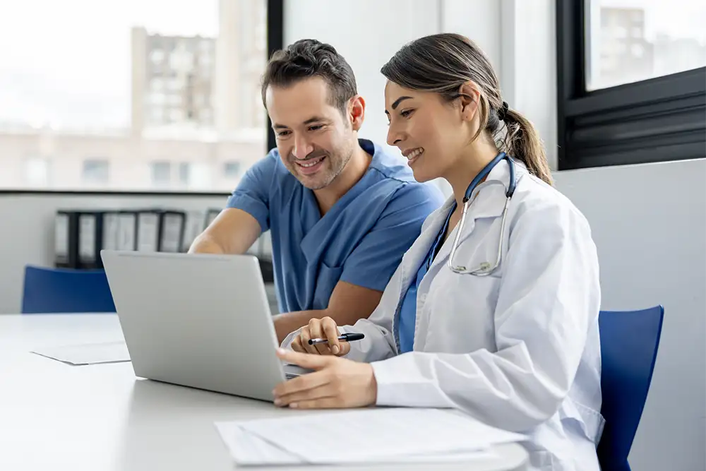 Two physicians sitting at a table smiling and looking at a laptop with papers in front of them